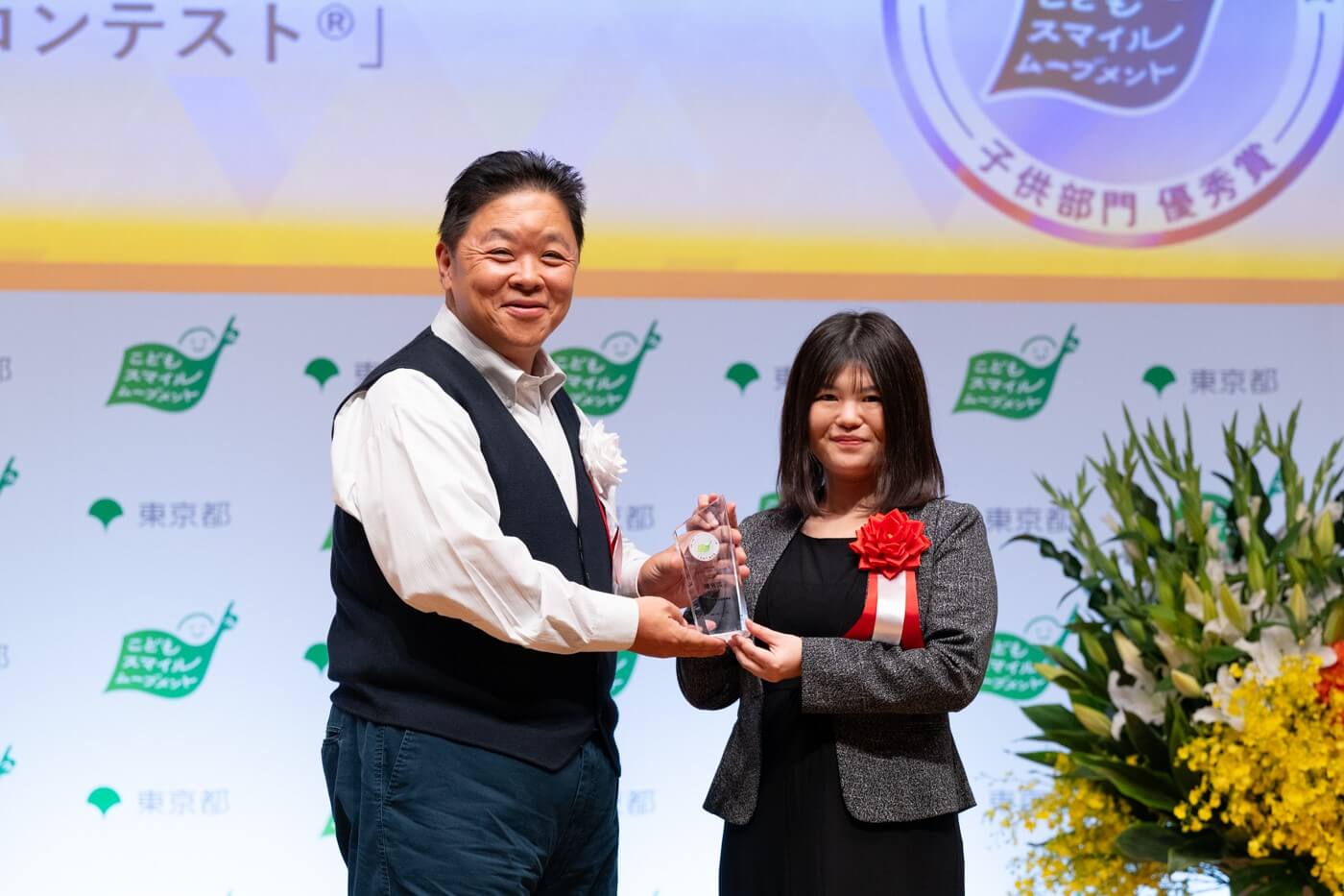 一般社団法人日本金融教育支援機構の代表者がこどもスマイルムーブメントアンバサダーの伊集院光さんから優秀賞の盾を受け取っている様子
