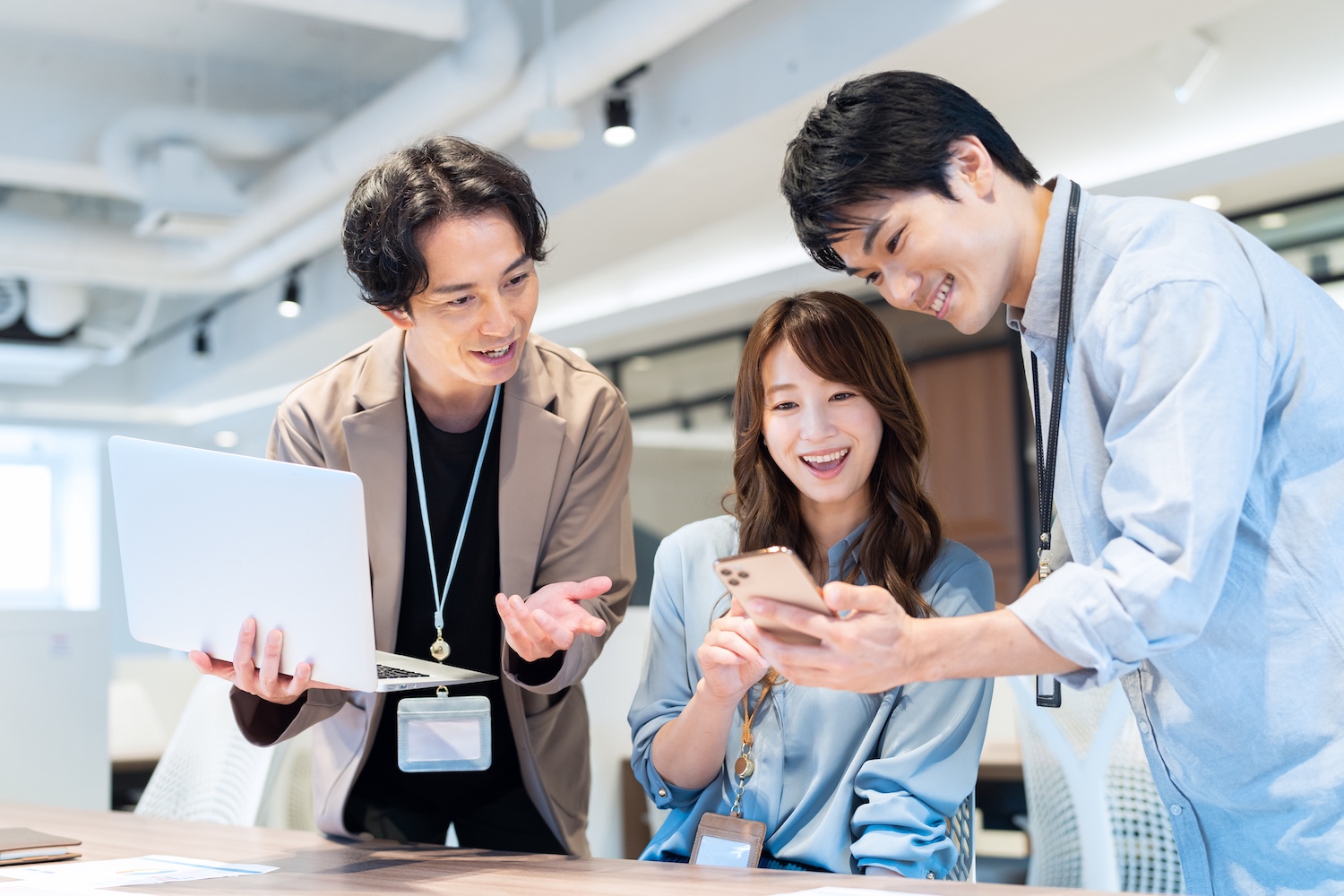 オフィスでパソコンとスマホを持ち話し合う男女3人の写真